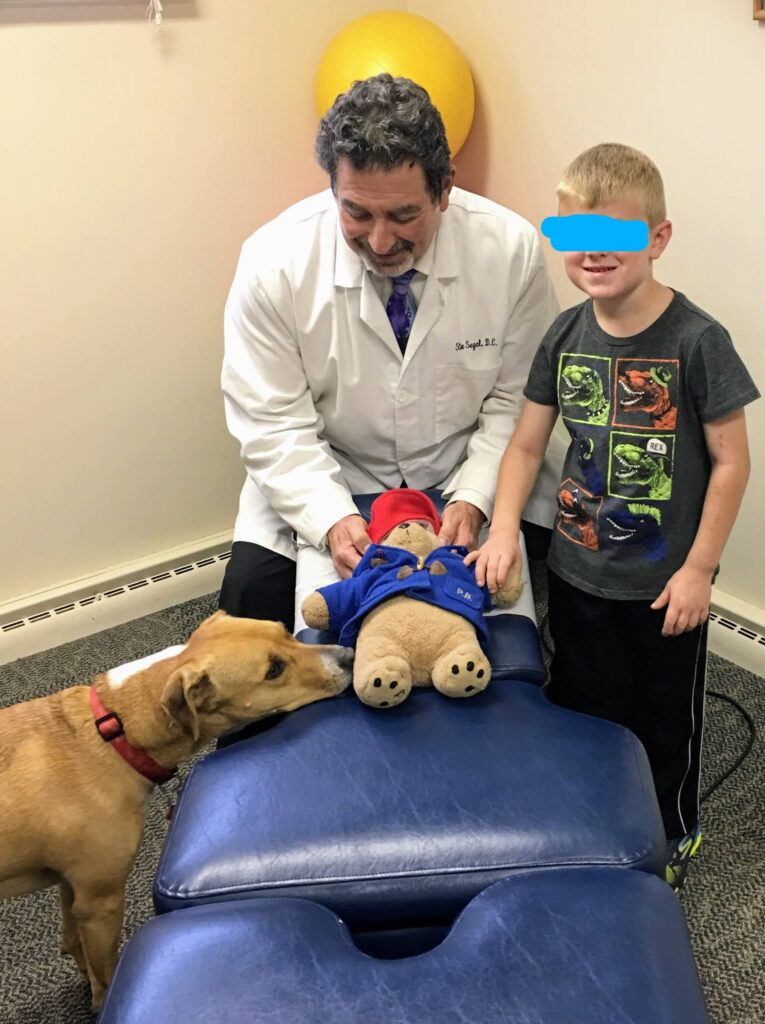 Dr. Steve Segal, Libertyville chiropractor, adjusting a child’s teddy bear while smiling with a young patient and friendly office dog nearby.